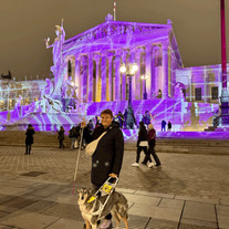 Lena Öllinger steht mit Assistenzhund vor dem lila beleuchteten Parlament in Wien