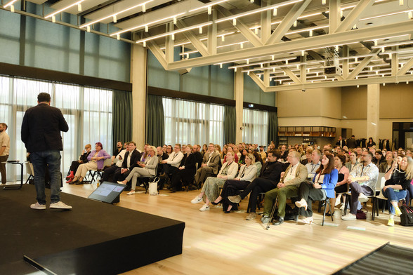 Eine Person auf einer Bühne spricht vor einem großen Publikum in einem hellen Saal