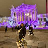 Lena Öllinger steht mit Assistenzhund vor dem lila beleuchteten Parlament in Wien