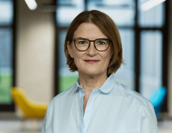 Eine Person mit Kinnlangen, hellbraunen Haaren und Brille steht vor einem Fenster. Sie trägt eine Brille und eine hellblaue Bluse.