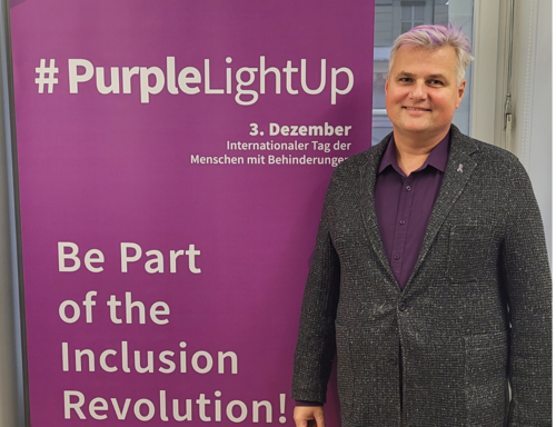 Robert Öllinger mit lila gefärbtem Haar, neben einem lila Roll-Up "#PurpleLightUp - 3. Dezember, Internationaler Tag der Menschen mit Behinderungen"