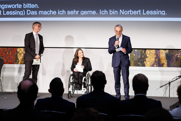 2 Männer auf der Bühne. In der Mitte die Moderatorin, die einen Rollstuhl nutzt.