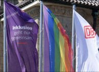 Es sind drei Flaggen abgebildet. Die linke Flagge ist lila mit dem Schriftzug Inklusion geht nur gemeinsam. Die Flagge in der Mitte ist mit den Regenbogenfarben. Die rechte Flagge ist weiß und hat das Logo der DB oben.