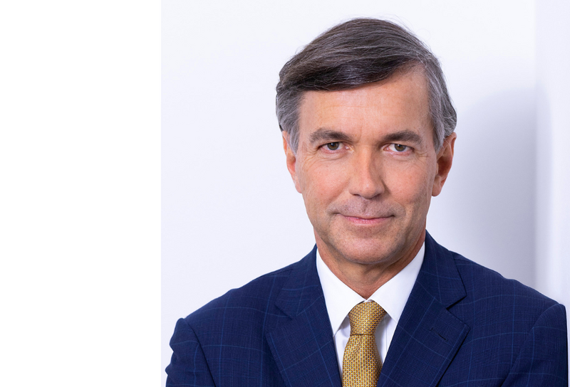 Die Person hat kurze graue Haare. Er trägt ein blaues Jackett mit einem weißen Hemd und einer gelben Krawatte.