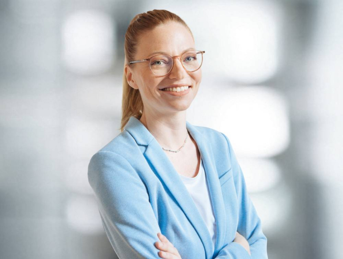 Porträt einer lächelnden Frau mit Brille, zurückgebundenen Haaren und hellblauem Blazer, die mit verschränkten Armen vor unscharfem Innenraum steht.