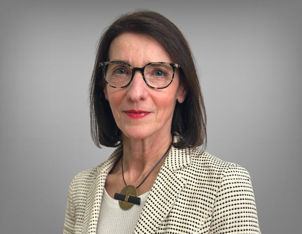 Portrait of Hilda Janik. She wears glasses and has shoulder-length black hair. She wears a necklace and a beige blazer with black dots. 