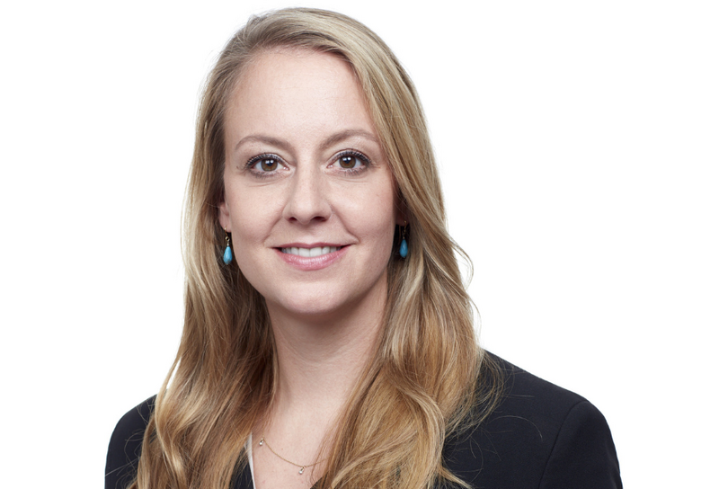 Die Person hat lange blonde Haare und Ohrringe. Sie trägt ein weißes Hemd und einen schwarzen Blazer. 