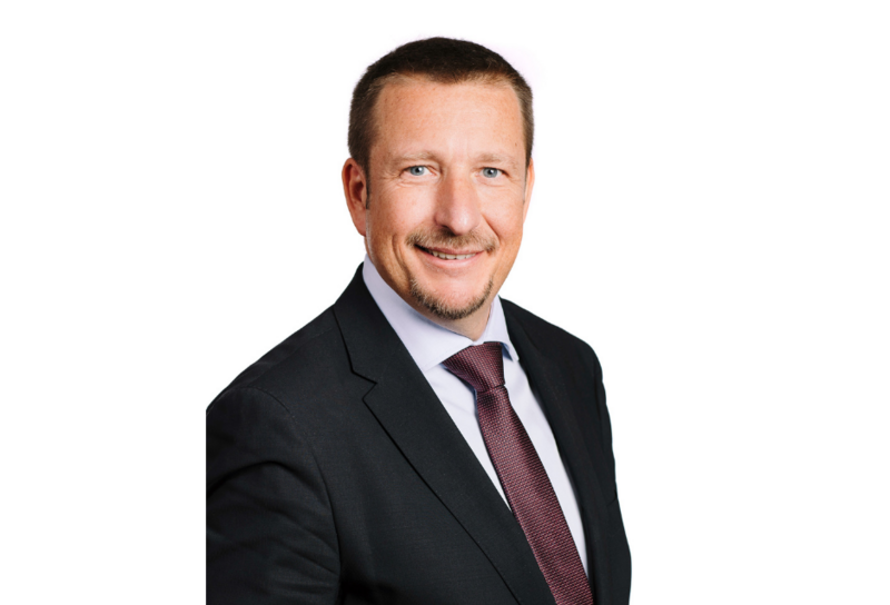Die Person hat kurze dunkle Haare und einen Bart. Er trägt ein Sakko mit weißem Hemd und einer roten Krawatte. 