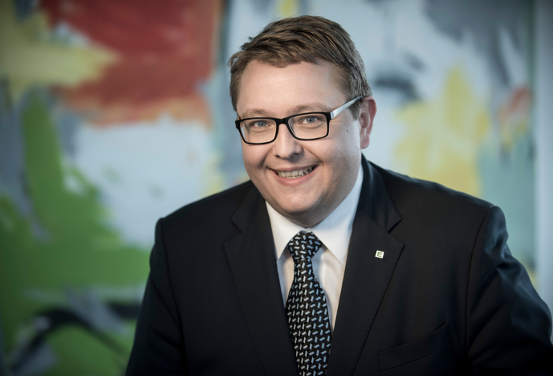 Die Person hat kurze braune Haare und eine Brille. Er trägt ein schwarzes Jackett mit weißem Hemd und einer gemusterten Krawatte. 