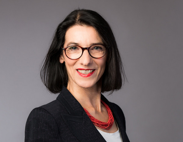 Portrait von Hilda Janik, weiblich gelesene Person mit halblangem schwarzem Haar, roter Kette und schwarzem Blazer