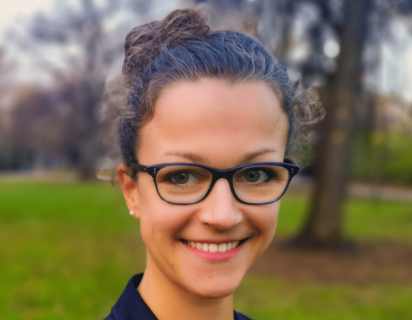 Person in einem Park mit zusammengebundenen, lockigen, dunkelbraunen Haaren. Sie trägt eine schwarze Brille und einen dunkelblauen Blazer. Sie lächelt in die Kamera.