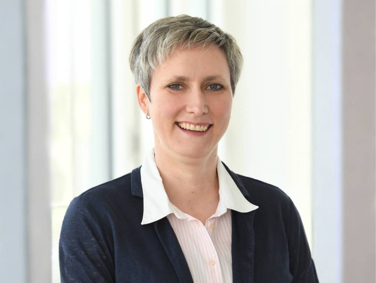Porträt einer lächelnden Frau mit kurzem Haar, dunkler Strickjacke und heller Bluse vor hellem, unscharfem Innenraum.
