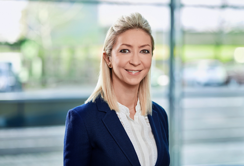 Die Person hat schulterlange blonde Haare. Sie trägt ein weißes Hemd mit einem dunkelblauen Blazer. 