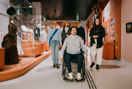 In einem Museum führt ein Guide, ein Rollstuhlnutzer mit kurzen braunen Haaren, eine Gruppe durch den Ausstellungsraum. An den Wänden hängen alte Fotografien. In einer Vitrine sind historische Kleider ausgestellt. Auf dem Boden sind Leitstreifen angebracht.