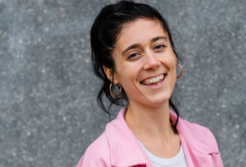 Die Person hat lange dunkle Haare, diese sind zu einem Zopf zusammen gebunden. Sie trägt eine rosa Jacke mit einem weißen T-Shirt. 