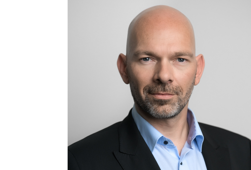 Person mit Glatze und Bart. Er trägt ein hellblaues Hemd mit einem schwarzen Blazer.
