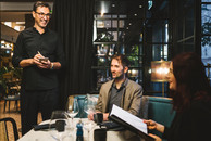 Ein Kellner mit Brille, dunklem Haar und schwarzem Hemd nimmt eine Bestellung auf. Am Tisch sitzt ein Mann im grauen Sakko und eine Frau mit rotem Haar. Der Tisch ist elegant gedeckt. Einer der beiden Gäste hat eine Sehbehinderung.