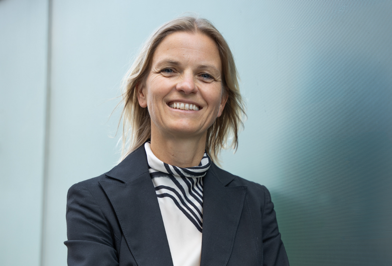 Die Person hat schulterlange brünette Haare. Sie trägt eine schwarzen Blazer mit einem weißen T-Shirt mit schwarzem Muster.
