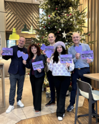 5 lila gekleidete Personen stehen vor einem Christbaum und halten lila Karten mit "PositivelyPurple" hoch