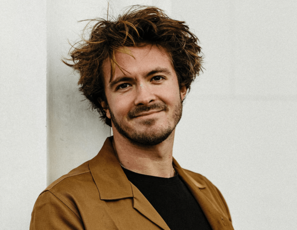 Person mit kurzen welligen braunen Haaren und Bart vor einer Hausmauer. Sie trägt ein braunes Sakko und ein schwarzes T-Shirt.