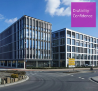 Zwei verglaste Büro Gebäude. Violettes Quadrat mit Text: DisAbility Confidence.