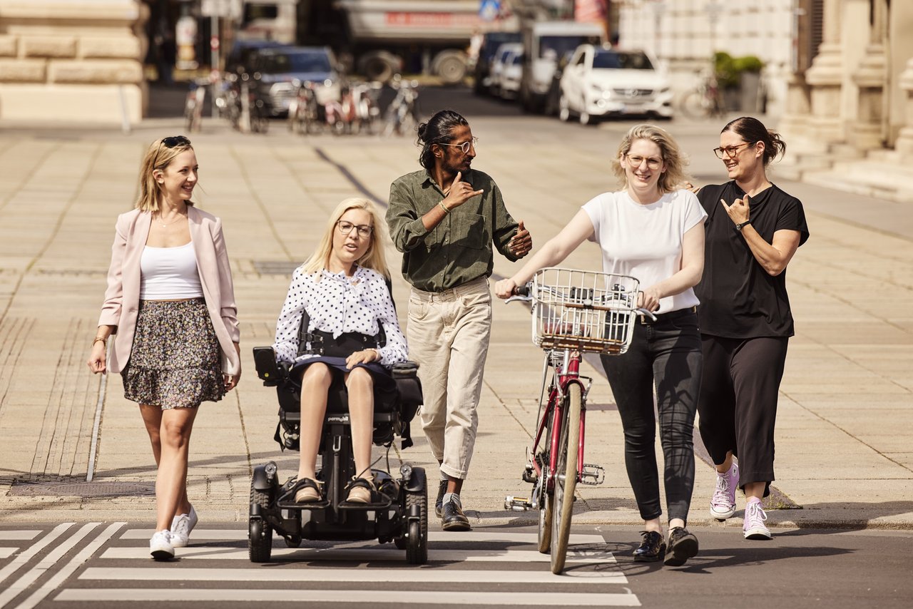 5 Personen mit sichtbaren und unsichtbaren Behinderungen überqueren die Straße. 