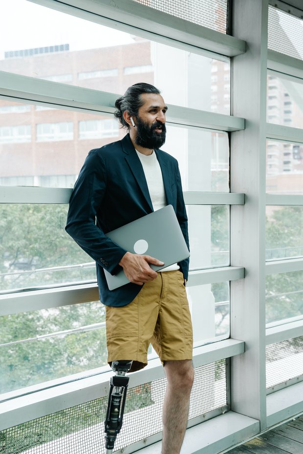 Mann mit Beinprothese und Laptop in der Hand steht bei einem Fenster 