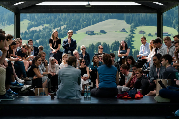 Fireside Chat: Teilnehmende sitzen dicht gedrängt im Kreis auf einer Bühne und hören zwei sprechenden Personen zu; dahinter Blick auf grüne Hänge und Wald.