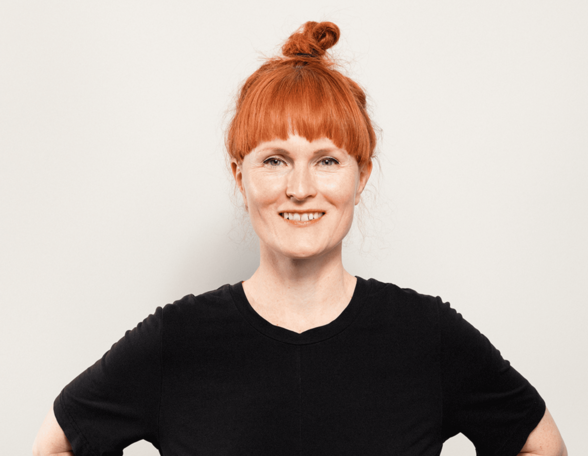 Person mit roten Haaren lächelt in die Kamera. Sie trägt die Haare in einem Dutt und hat ein schwarzes Kleid an.