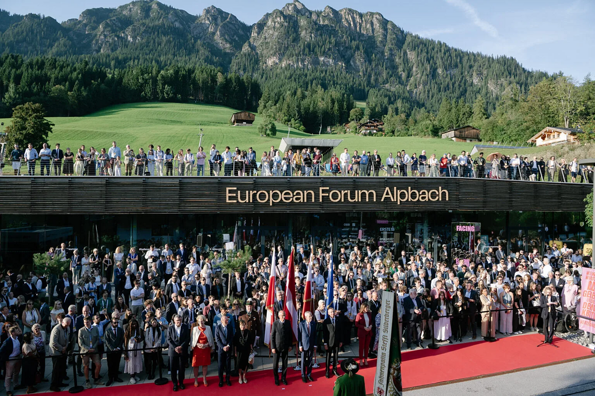 Große Menschenmenge vor einem Veranstaltungsgebäude mit der Aufschrift „European Forum Alpbach“, davor roter Teppich und mehrere Flaggen; im Hintergrund grüne Wiesen und Berge.