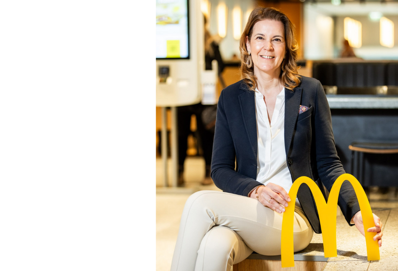 Die Person hat schulterlange braune Haare und Ohrringe. Sie trägt eine weiße Bluse mit einem schwarzen Blazer und einer beigen Hose. Sie sitzt und hält mit beiden Händen das Logo von McDonalds.