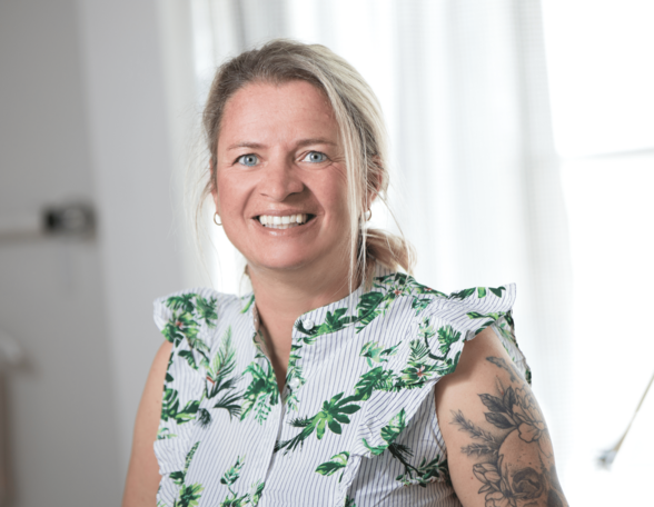 Person mit blonden Haaren, zu einem Zopf gebunden, lächelt in die Kamera. Sie hat ein Tattoo am linken Oberarm und trägt eine gemusterte Bluse