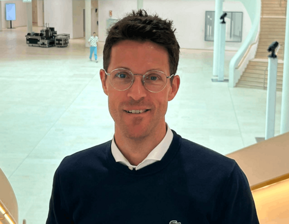 Person mit kurzen braunen Haaren und Brille steht in einer Eingangshalle. Sie trägt eine Brille, ein weißes Hemd und einen dunkelblauen Pullover.