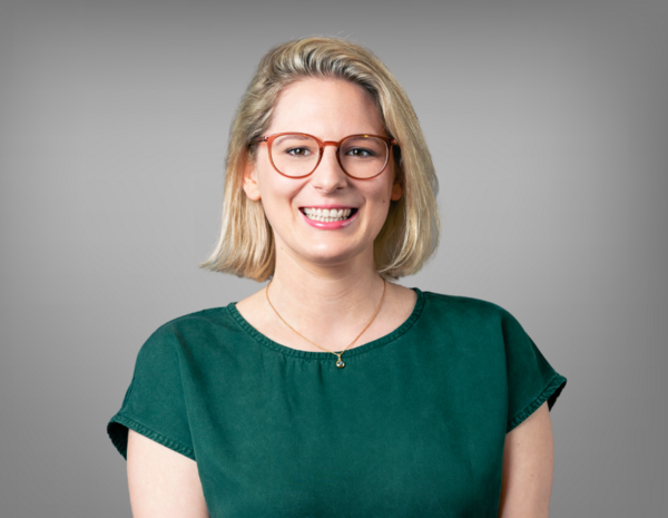 Portrait von Julia Diexer, weiblich gelesene Person mit schulterlangem blonden Haar und roter Brille, in weißer Bluse mit dunkelblauer Weste