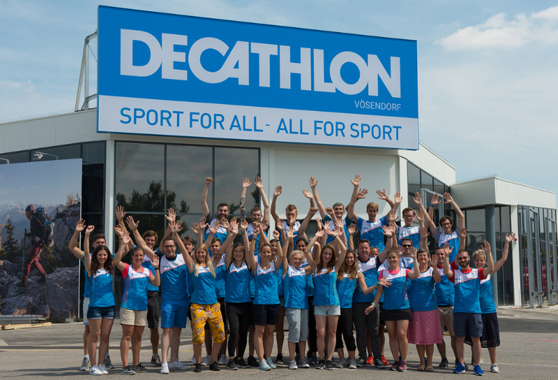 Eine Gruppe von Personen steht mit ausgestreckten Armen in die Luft vor einer Decathlon Filiale.