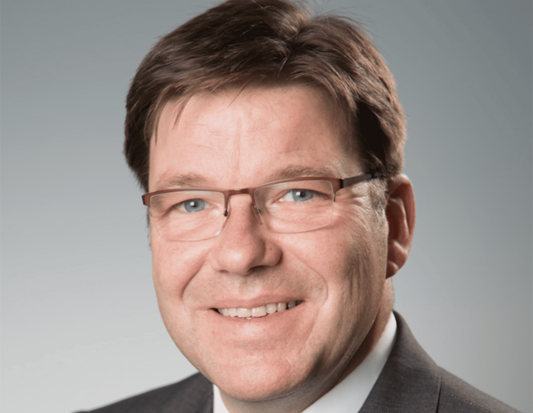 Person mit dunklebraunen Haaren und Brille in grauem Anzug mit blauer Kravatte vor grauem Hintergrund.