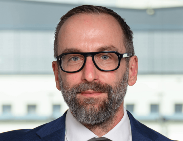 Person mit kurzen Haaren, graumeliertem 3-Tage-Bart und Brille. Sie trägt einen dunklen Anzug mit Krawatte und ein weißes Hemd.