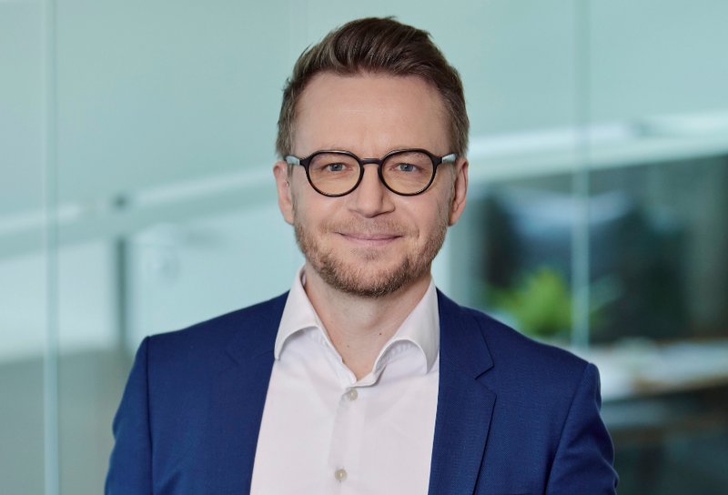 Die Person hat kurze braune Haar, einen Bart und eine Brille. Er trägt ein blaues Jackett mit einem weißen Hemd.