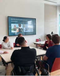 6 Personen sitzen rund um einen Tisch und unterhalten sich. Im Hintergrund ein Fernseher mit eine Präsentationsfolie von myAbility
