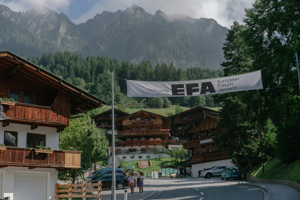 Straßenszene mit alpinen Häusern und Bergkulisse; über der Straße hängt ein Banner mit dem Text „Welcome to the EFA European Forum Alpbach“.