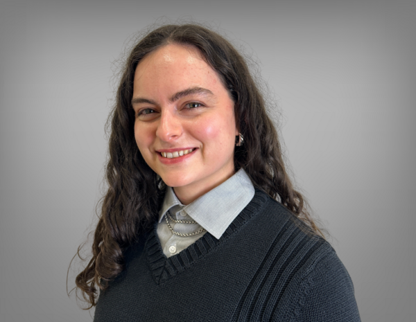 Laura hat dunkles, lockiges schulterlanges Haar. Sie trägt ein helles Hemd mit einem dunklen Pullover darüber.