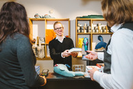 Zwei Personen stehen an der Kasse eines Geschäfts. Der Verkäufer, ein Mann mit kurzen blonden Haaren und Brille, nimmt die Ware entgegen. Er lächelt freundlich.