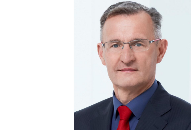 Person mit kurzem grauen Haar blickt in die Kamera. Sie trägt eine Brille mit dünnem Alurand, ein dunkles Sakko, ein blaues Hemd und eine rote Krawatte.