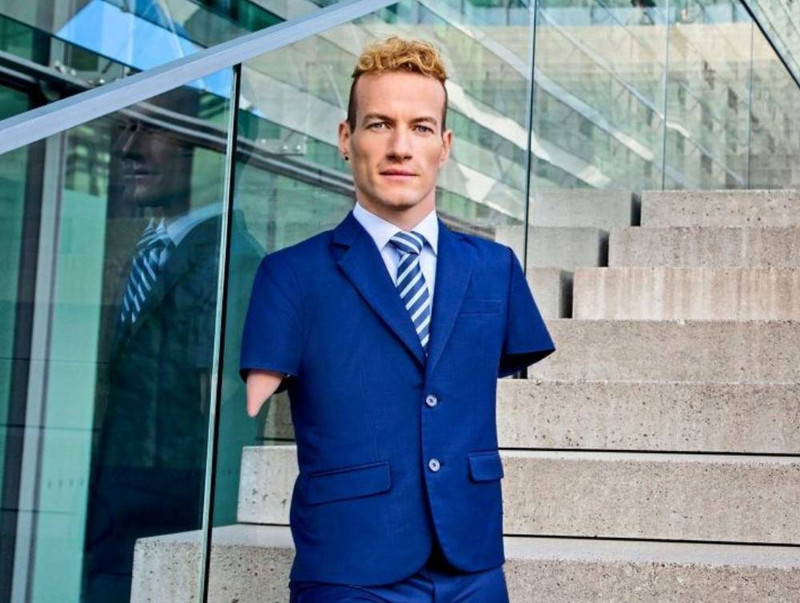 Janis McDavid hat blonde kurze Locken und trägt einen dunkelblauen Anzug und eine gestreifte Krawatte. Er hat keine Arme und ist auf einer Treppe abgelichtet. Im Hintergrund ein gläsernes Gebäude