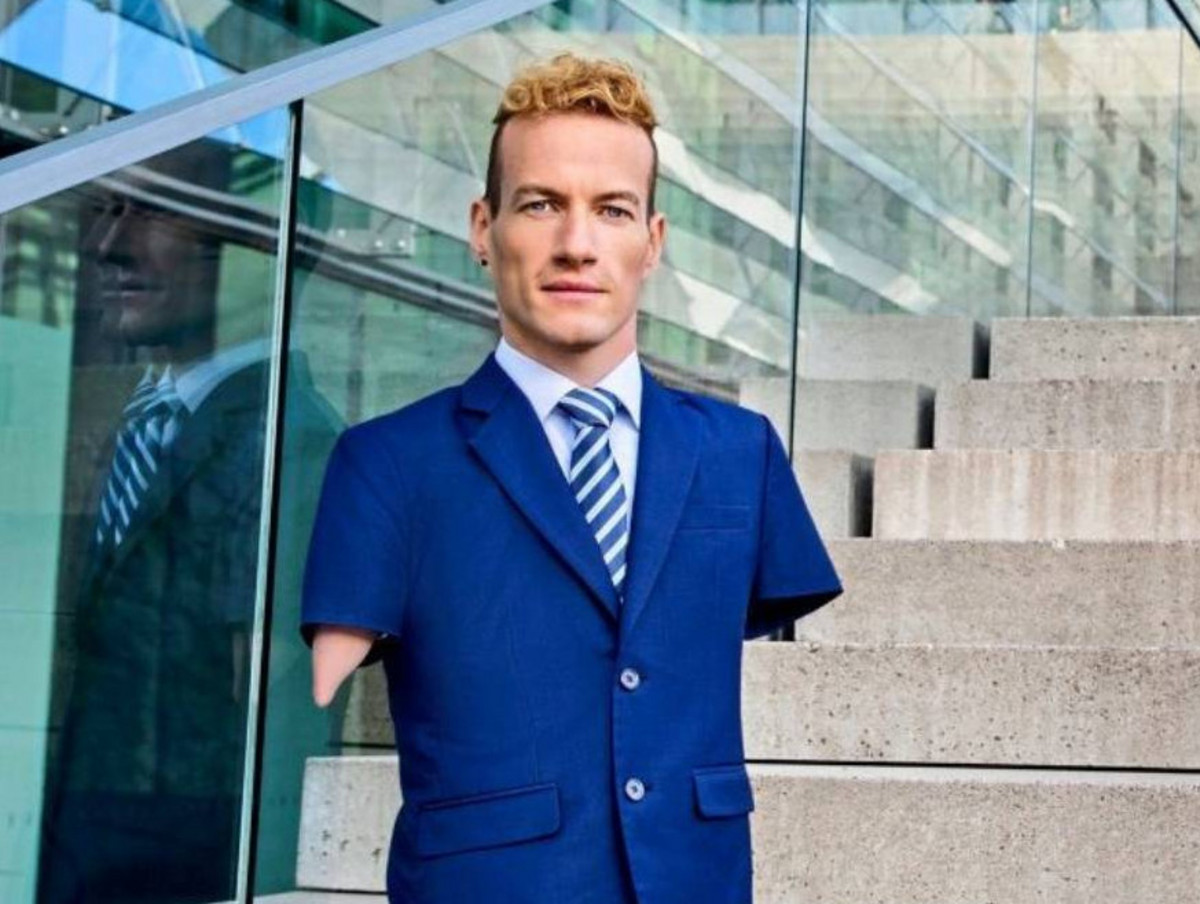 Janis McDavid hat blonde kurze Locken und trägt einen dunkelblauen Anzug und eine gestreifte Krawatte. Er hat keine Arme und ist auf einer Treppe abgelichtet. Im Hintergrund ein gläsernes Gebäude