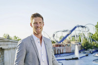 Nicolas Mack trägt ein graues Sakko und hat kurzes braunes Haar. Er steht vor dem Gelände des Europa-Parks. Im Hintergrund ist eine Achterbahn zu sehen.