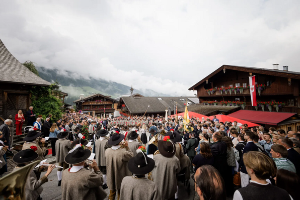 Menschenmenge in einem Dorfplatz um eine Musikkapelle in Tracht, neben einer Bühne mit Redner:innen; an den Häusern hängen rot-weiß-rote Fahnen.