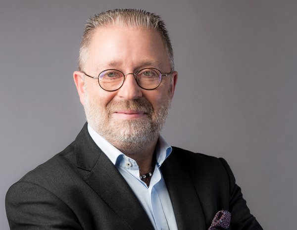 Portrait of Erik Schäfer, male person with gray hair and beard. He wears glasses, a light blue shirt and a black jacket.
