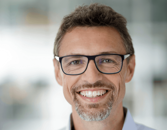 Person mit kurzen Haaren, graumeliertem Bart und Brille lächelt in die Kamera. Sie trägt ein blaues Hemd.
