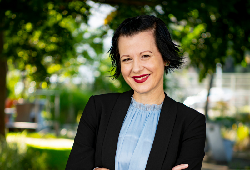 Die Person kurze schwarze Haare und trägt Lippenstift. Sie trägt ein blaues T-Shirt und einen schwarzen Blazer.